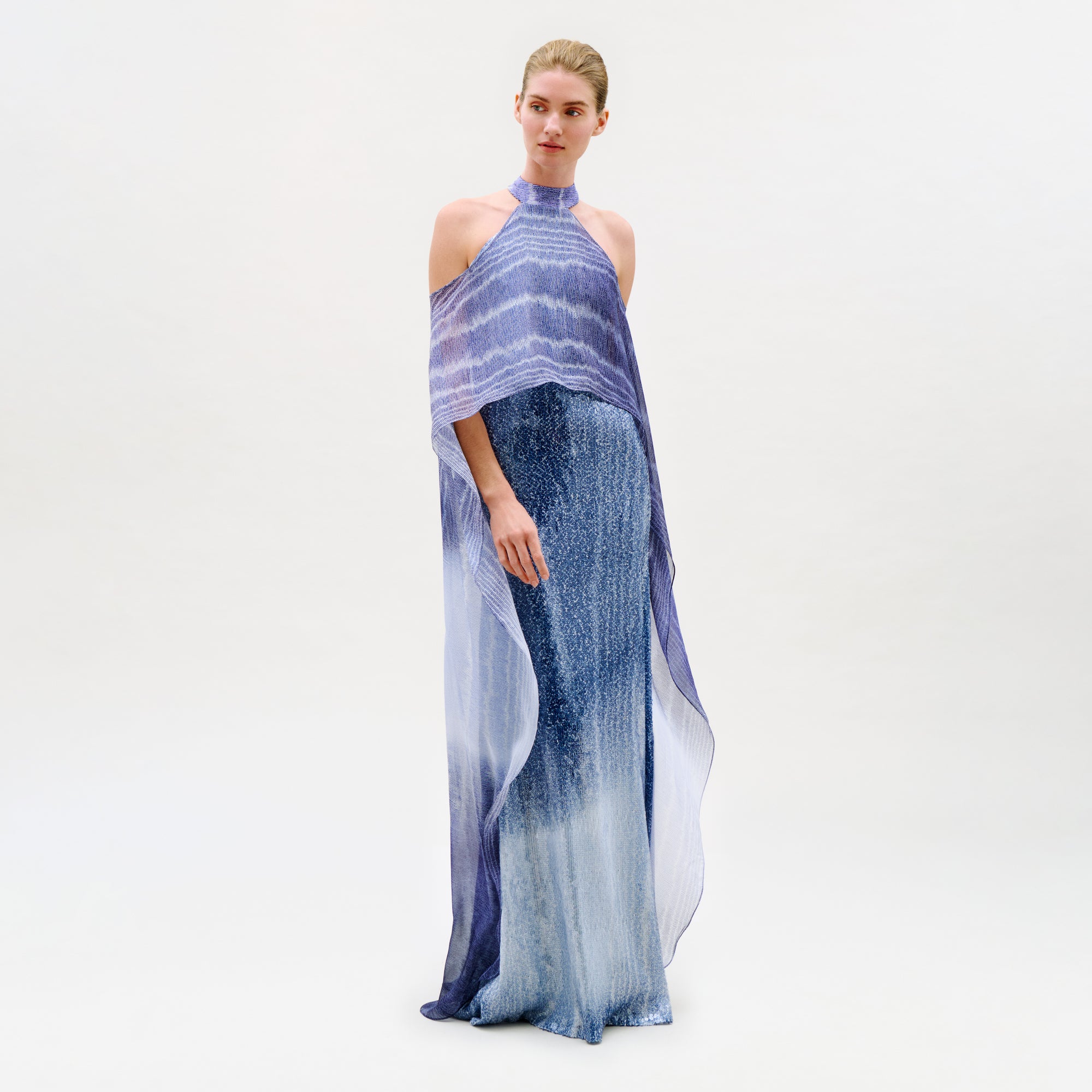 A woman stands against a plain white background wearing the Serenela Dress—a floor-length, sleeveless maxi dress in a blue-purple gradient with a halter neckline and sheer overlay cape detail, expertly crafted from luxurious Italian fabric.