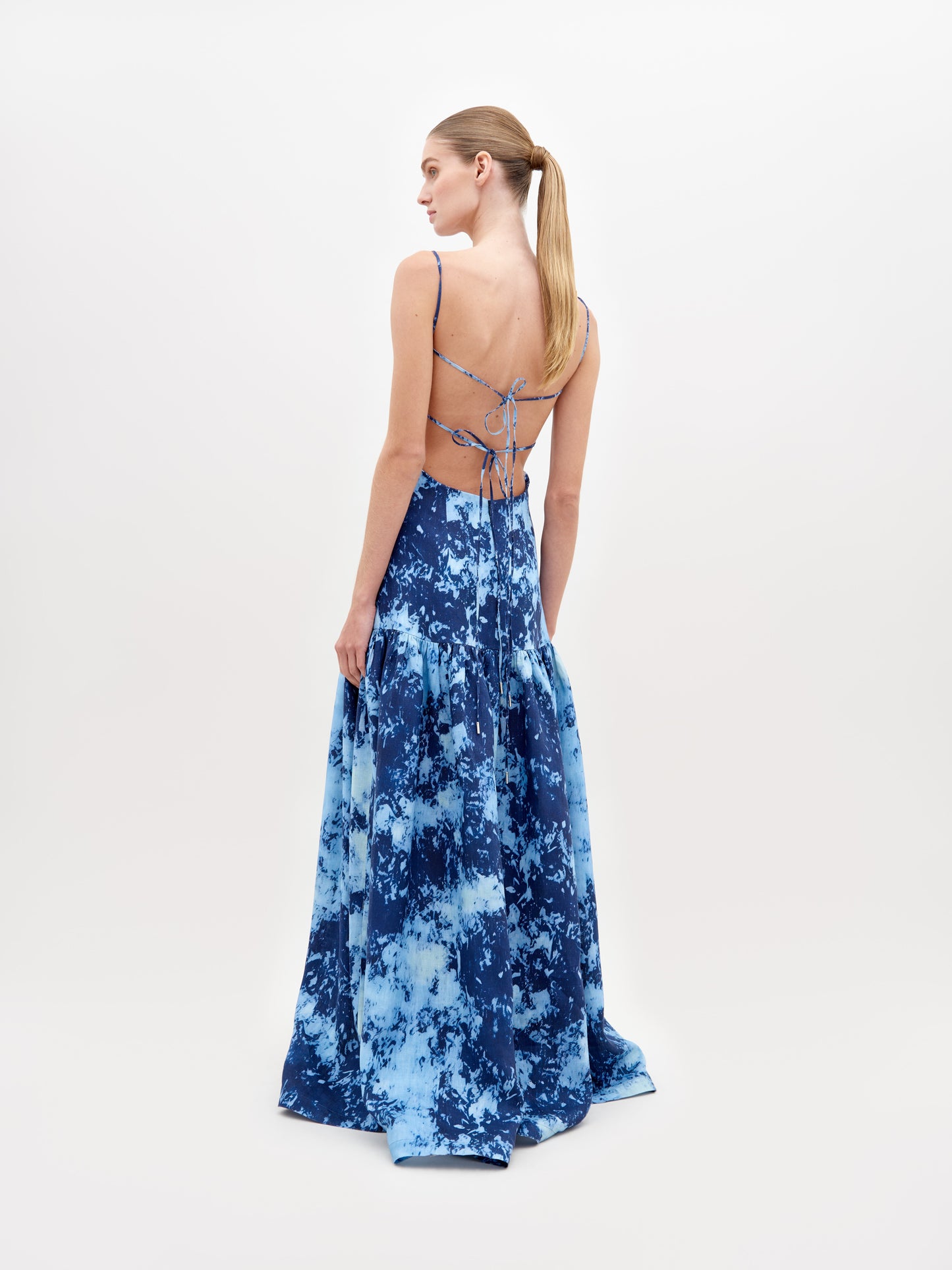 A woman stands wearing a long, blue, tie-dye sleeveless dress with thin straps and side cutouts, posing against a plain white background. The Shannon Dress Indigo is available for pre-order now and will ship by November 15th, 2024.