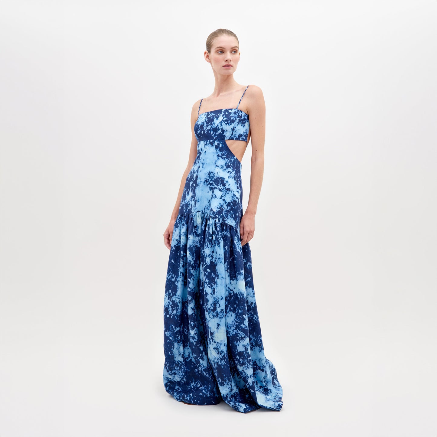 A woman stands wearing a long, blue, tie-dye sleeveless dress with thin straps and side cutouts, posing against a plain white background. The Shannon Dress Indigo is available for pre-order now and will ship by November 15th, 2024.