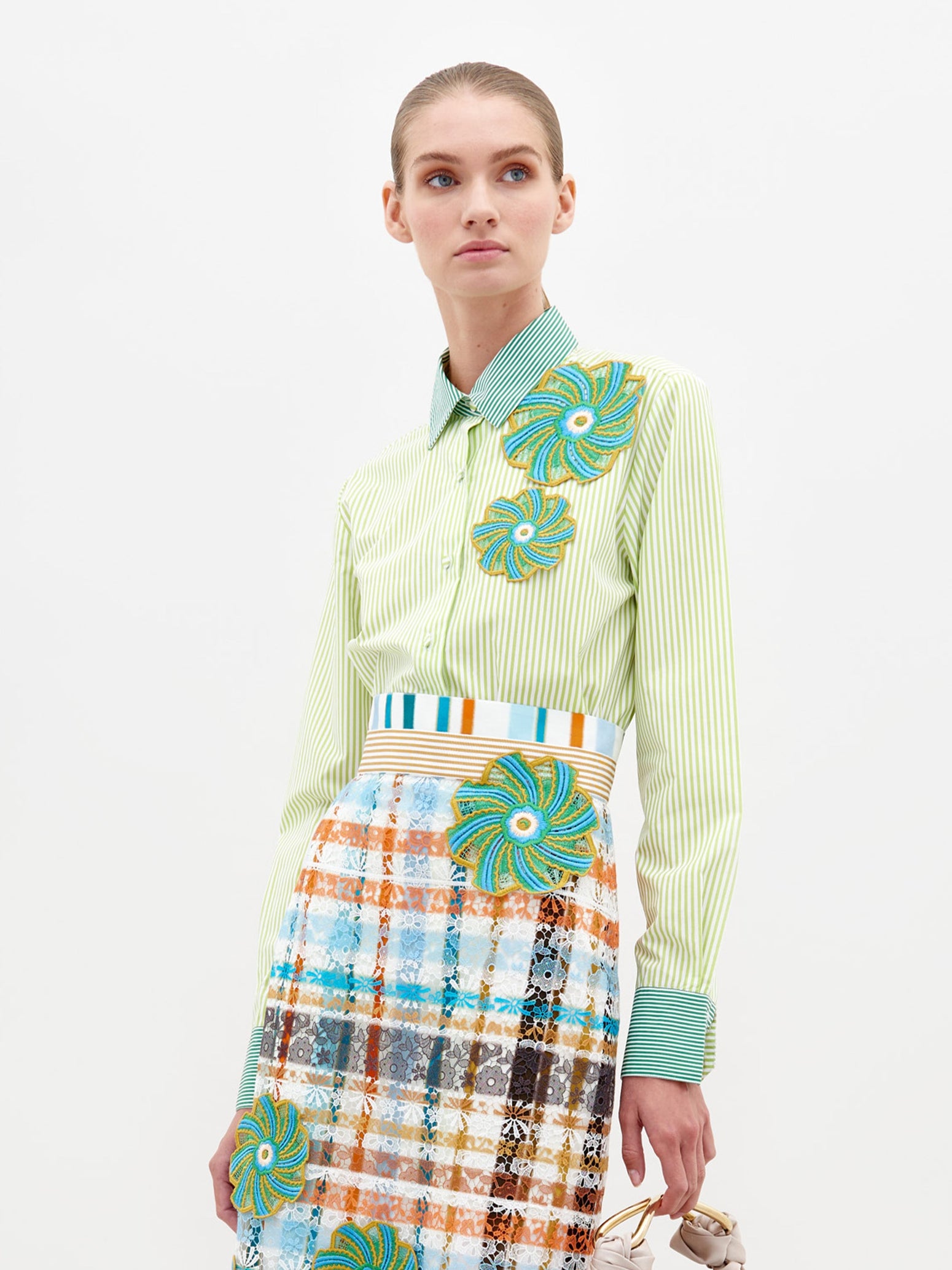 A woman stands against a plain background wearing the Shao Blouse Green Stripes, adorned with colorful floral embellishments, and a multicolored plaid skirt. She holds a small item in her left hand, now available for pre-order and expected to ship by February 15th, 2025.