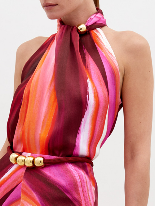 A woman stands against a plain background, wearing a halter-neck dress with a multicolored pattern of pink, orange, and red hues, accessorized with a gold belt buckle. Pre-order the Shela Blouse Magenta Abstract Waves now to ensure delivery by November 15th, 2024.