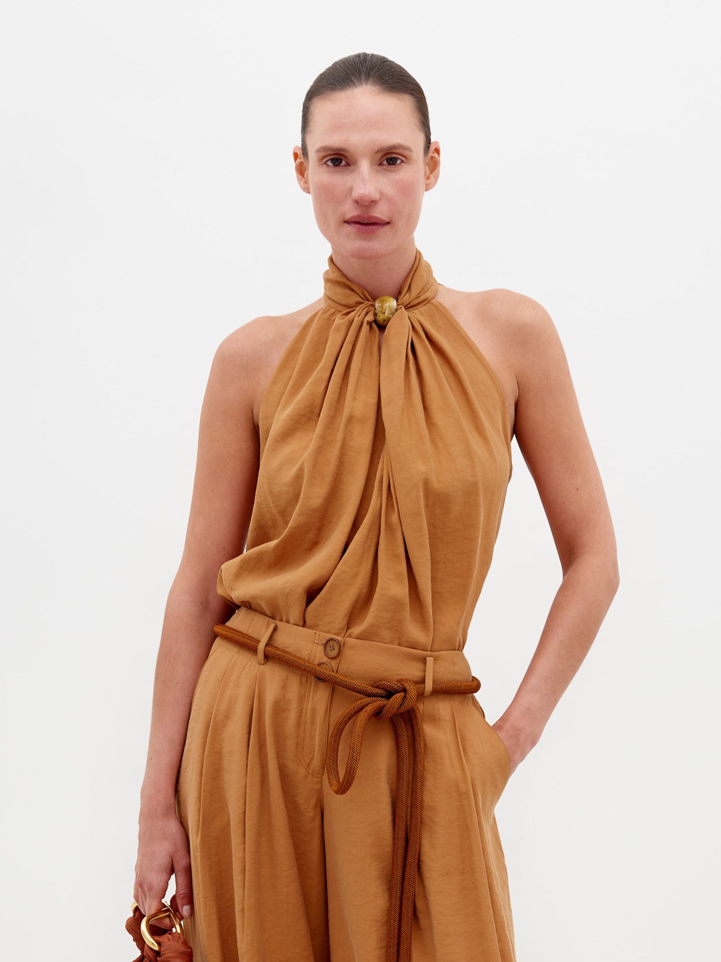 A person wearing the Shela Blouse Camel sleeveless high-neck top with matching wide-leg trousers, accessorized with a belt and holding a small object, stands against a plain white background. Pre-order now to ensure it ships by February 15th, 2025.