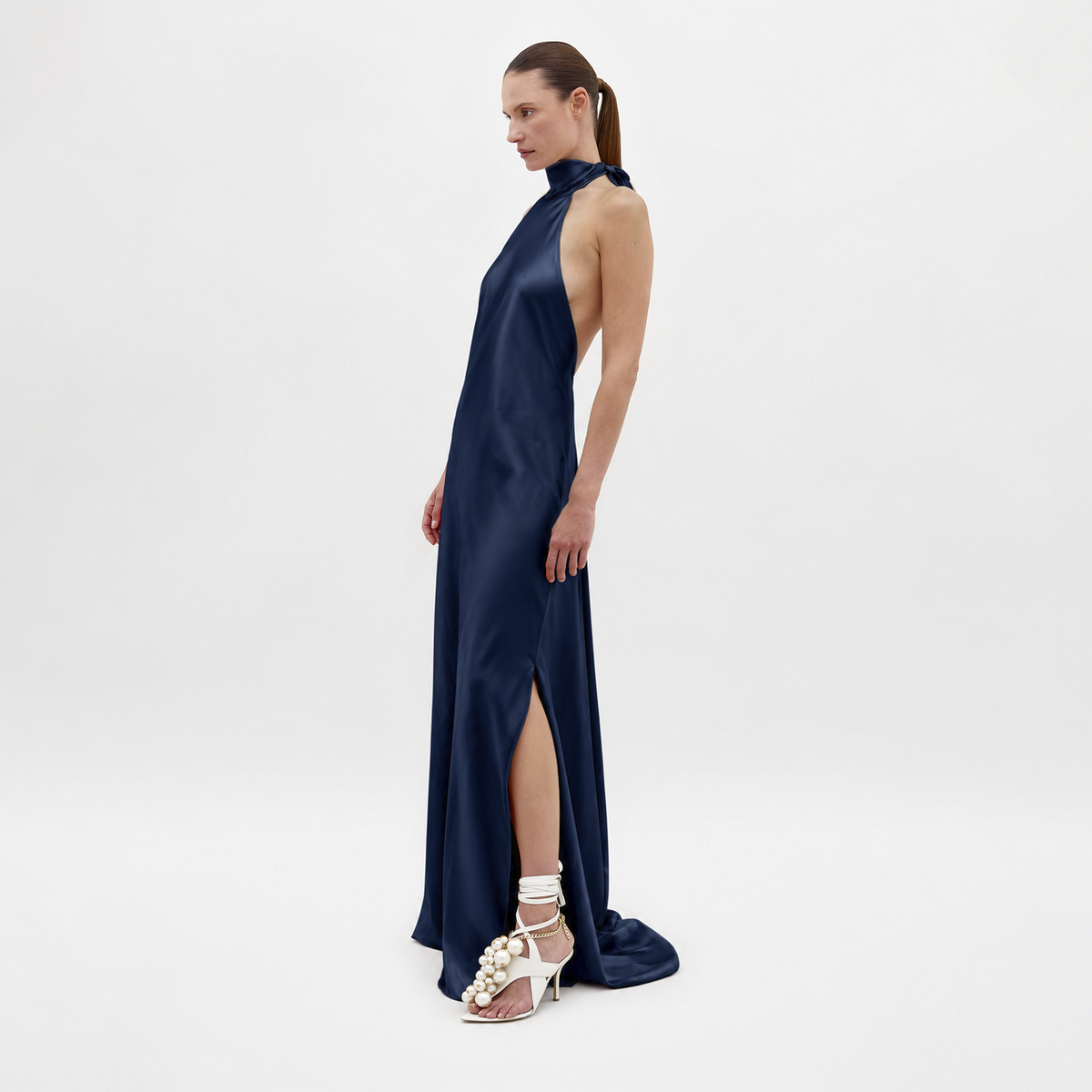 The Sherry Dress Navy is a floor-length halter-neck dress in navy with an open back and a high leg slit, paired with white embellished heeled sandals.