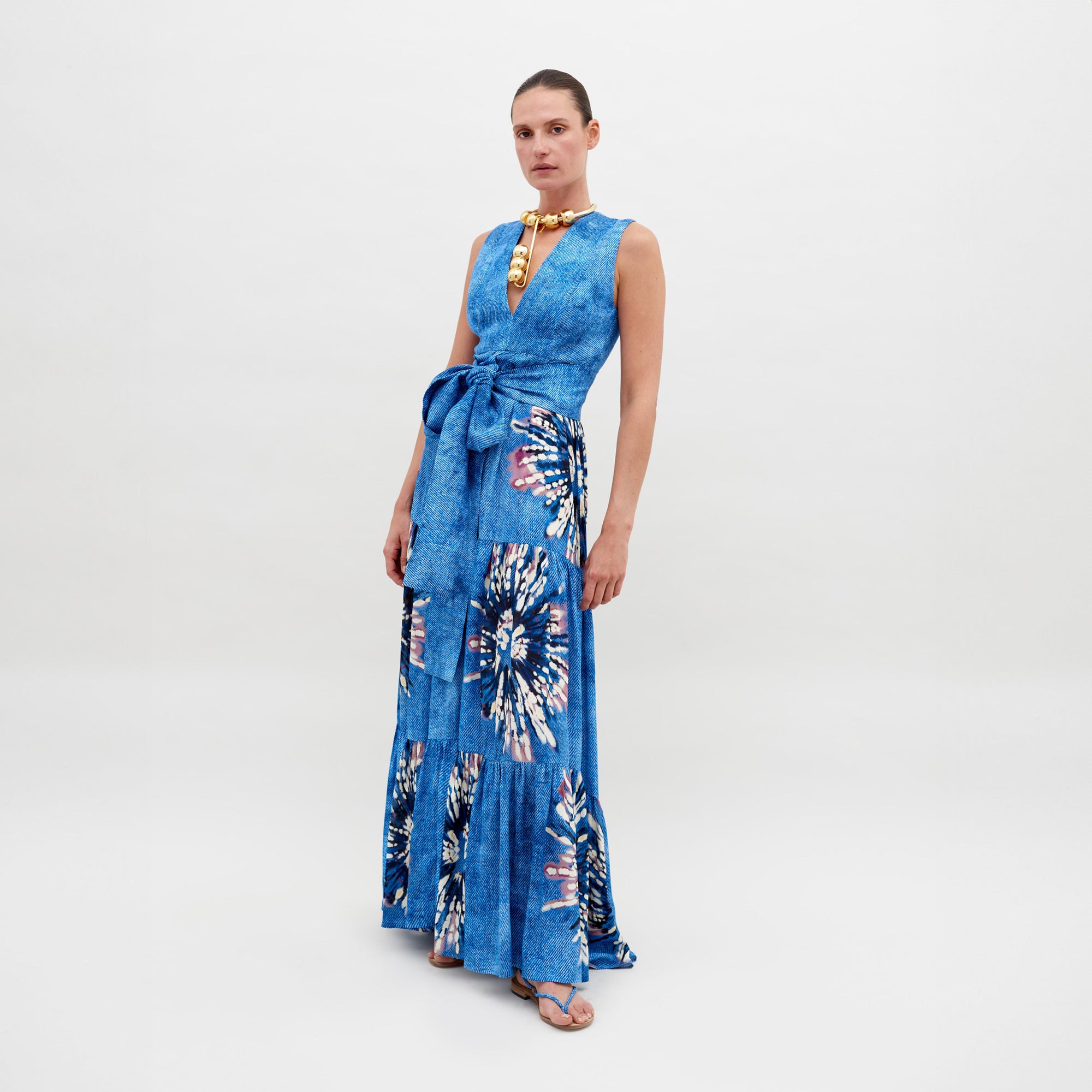 A woman stands wearing a Shirley Dress Aciano Flower with floral patterns at the bottom, a gold-colored choker, and flat sandals. She has her hair pulled back and is posing against a plain white background. Pre-order now for a ship date in November 2024.