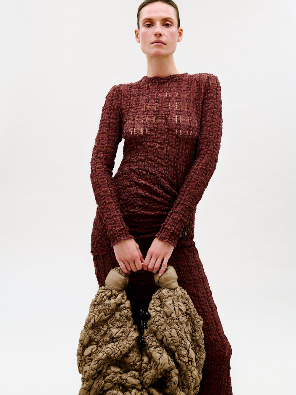 A model wearing a textured, long-sleeved burgundy dress and holding a chunky, crinkled taupe-brown bag against a plain light gray background.