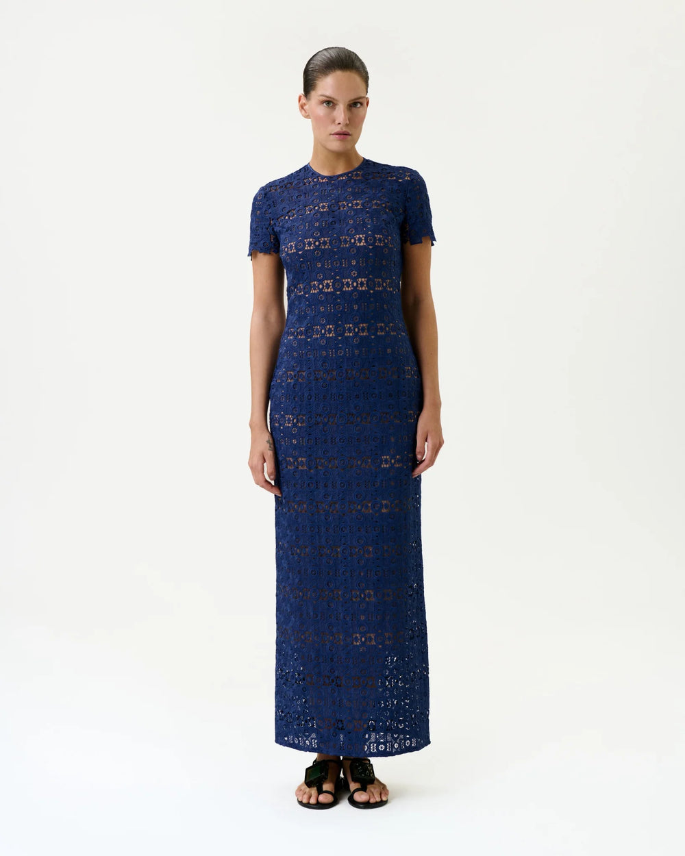 A woman stands against a plain white background wearing the Taiana Dress—a fitted, short-sleeve, ankle-length navy blue lace maxi made in Colombia with a high neckline—paired with black sandals.