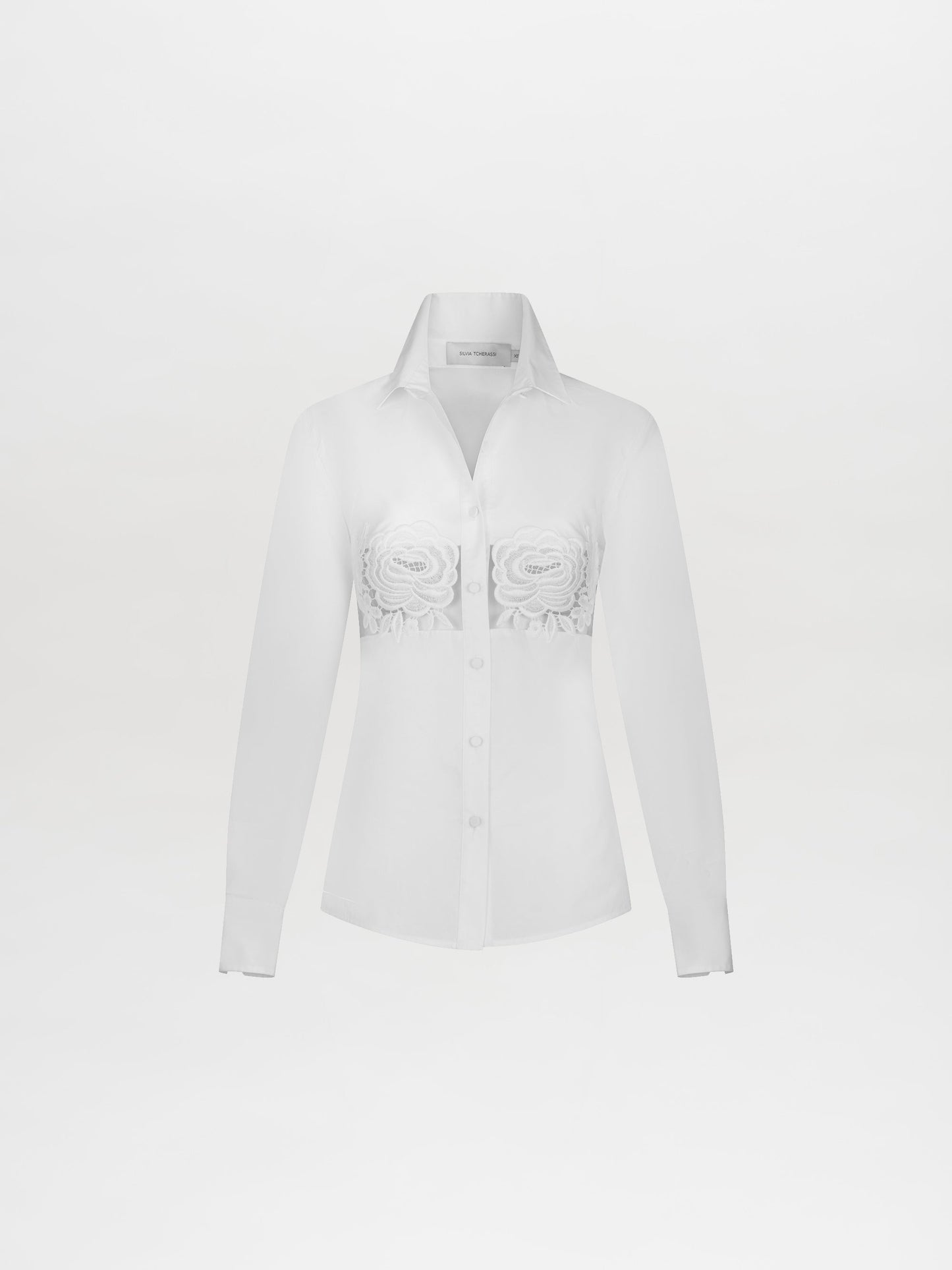 A person wearing the Sigrid Blouse White, a long-sleeved top featuring floral cut-outs, paired with high-waisted white pants with a floral belt, stands against a plain white background. Ready for pre-order, mark your calendars for February 15th, 2025, the anticipated ship-by date.