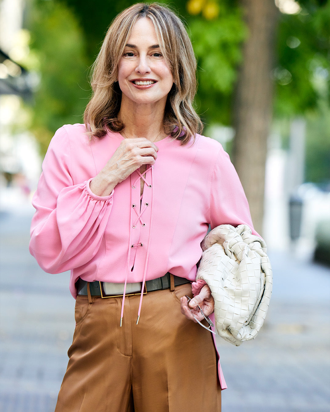 Woman in a pink blouse and tan pants, holding a woven bag, stands outdoors on a sunny day.