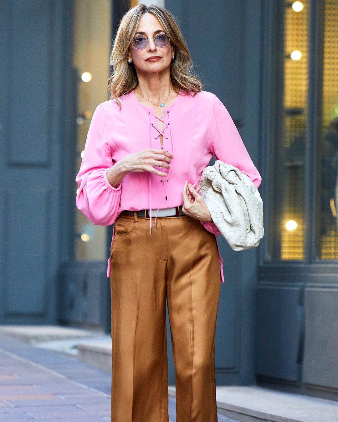 A person wearing a pink blouse and gold pants stands on a city street, holding a large, light-colored bag.