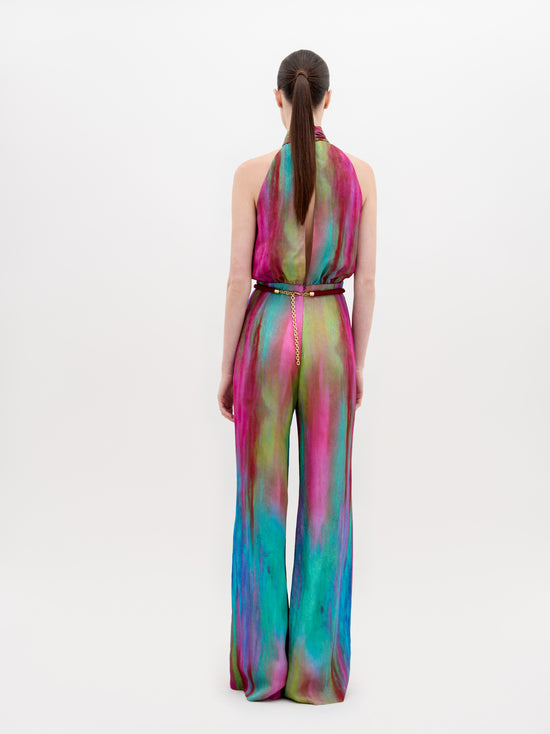 A woman stands wearing a vibrant, multicolored Siv Jumpsuit Watercolor with a belt. She has her hair pulled back and is posing against a plain white background. This eye-catching outfit is available for pre-order now, with a ship by date of November 15th, 2024.