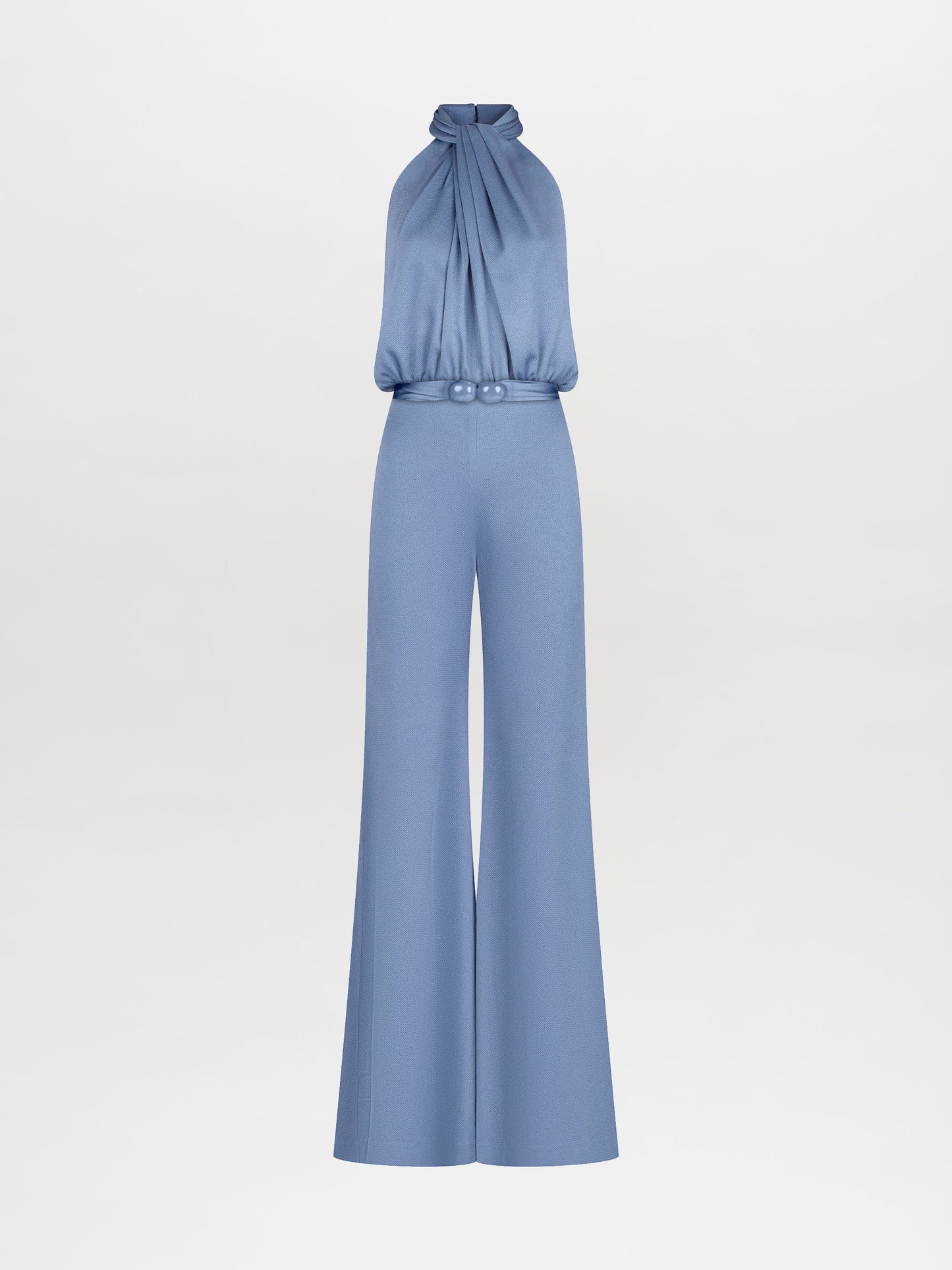 A person wearing the Siv Jumpsuit Periwinkle, featuring a sleeveless, light blue, halterneck design with wide-leg pants and a belted waist, stands against a plain white background. Pre-order now to ensure it ships by February 15th, 2025.