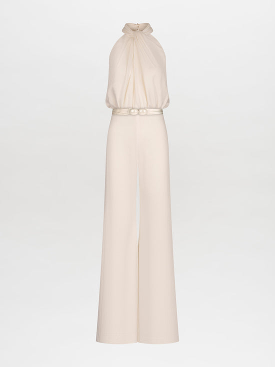 A woman stands against a plain background, wearing the Siv Jumpsuit White—a sleeveless halter jumpsuit featuring a beaded embellishment at the waist. This stunning piece is now available for pre-order and is set to ship by February 15th, 2025.