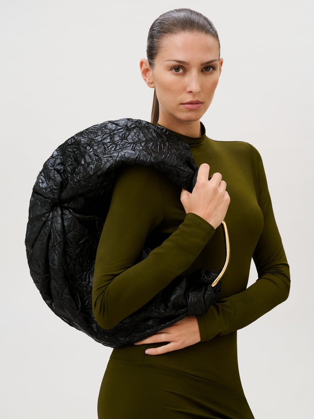 A person in an olive green, long-sleeved dress stands holding the Sofia Bag (Maxi) with a gold-toned handle over one shoulder, looking directly at the camera against a plain light background.