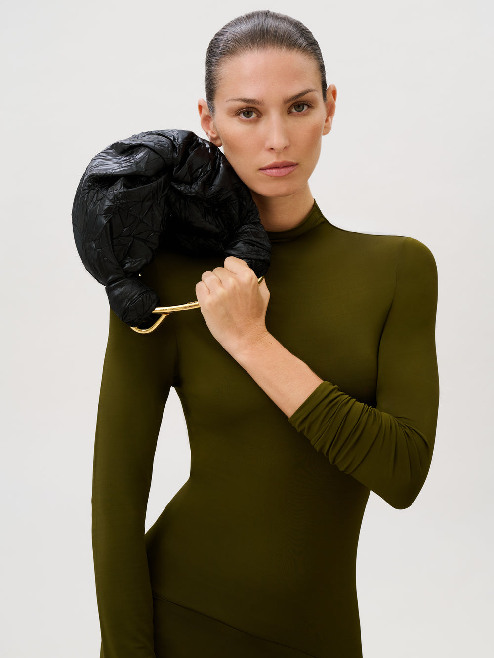 A woman in a fitted olive-green dress holds the Sofia Bag (Small), crafted from Italian fabric with a gold-toned handle, on her shoulder while standing against a plain light background and looking directly at the camera with a serious expression.