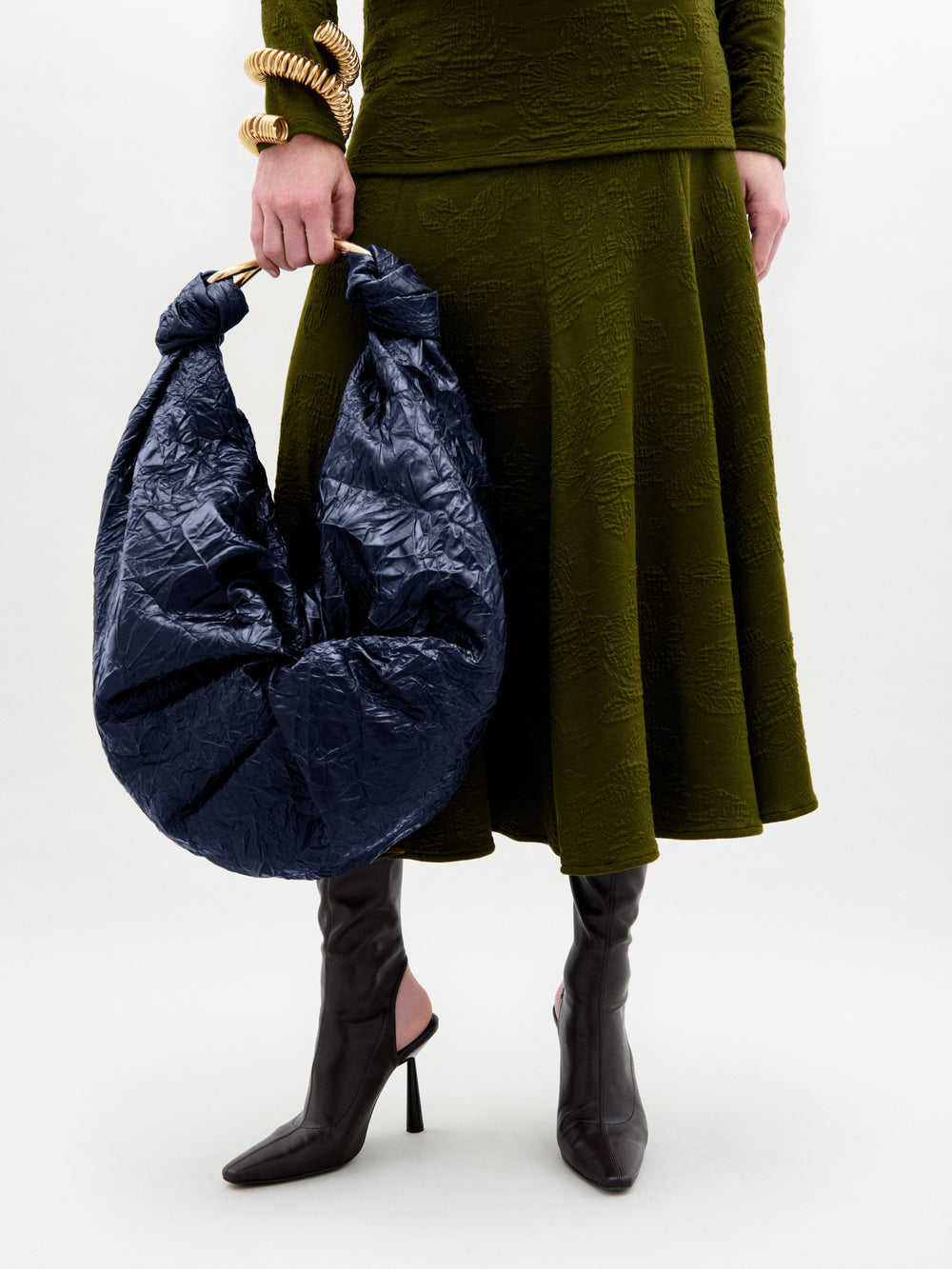 A person wearing a textured olive green skirt and top, black heeled boots, chunky gold bracelets, and holding the Sofia Bag (Maxi)—a large bag crafted from crinkled navy blue Italian fabric with a gold-toned handle.
