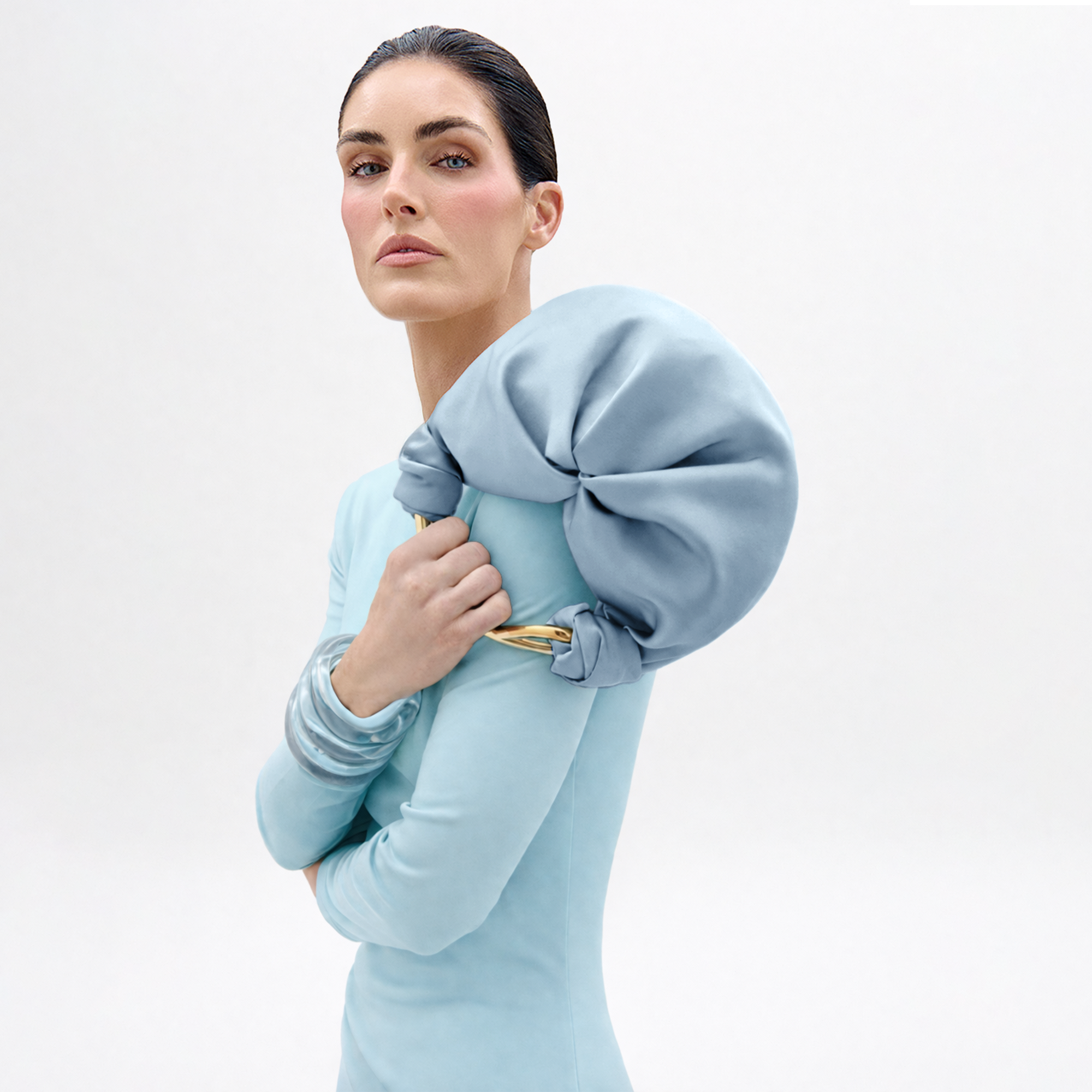 A woman with slicked-back dark hair and subtle pink makeup poses against a white background, holding the Sofia Bag (Small)—a soft, light blue clutch with gold accents—over her shoulder while wearing a pale blue outfit.