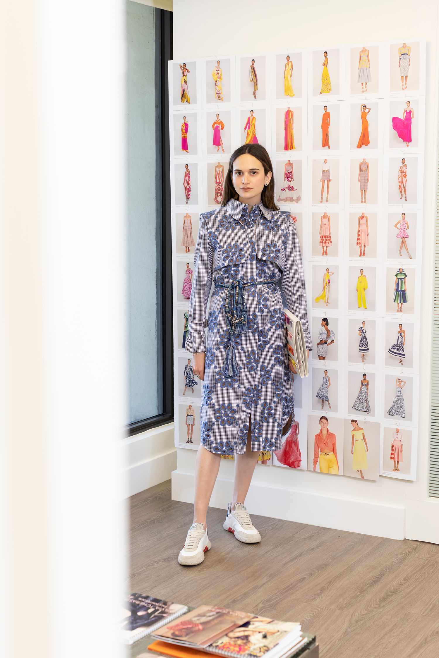 A woman stands in a room by a collage of fashion illustrations, wearing a patterned dress and holding magazines.