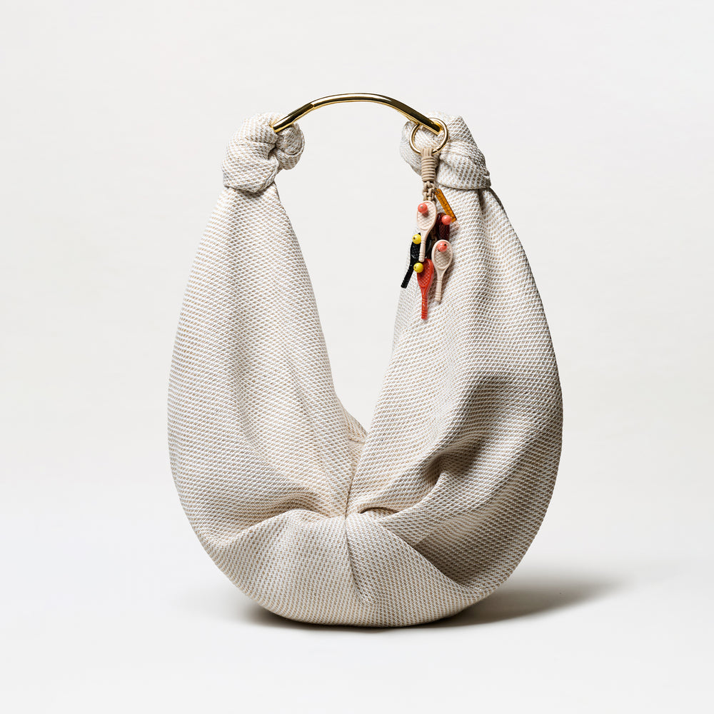 The Tournament Charm is a cream-colored, textured fabric hobo bag with a gold metal handle and several small, colorful keychain charms hanging from one side, displayed against a plain white background.