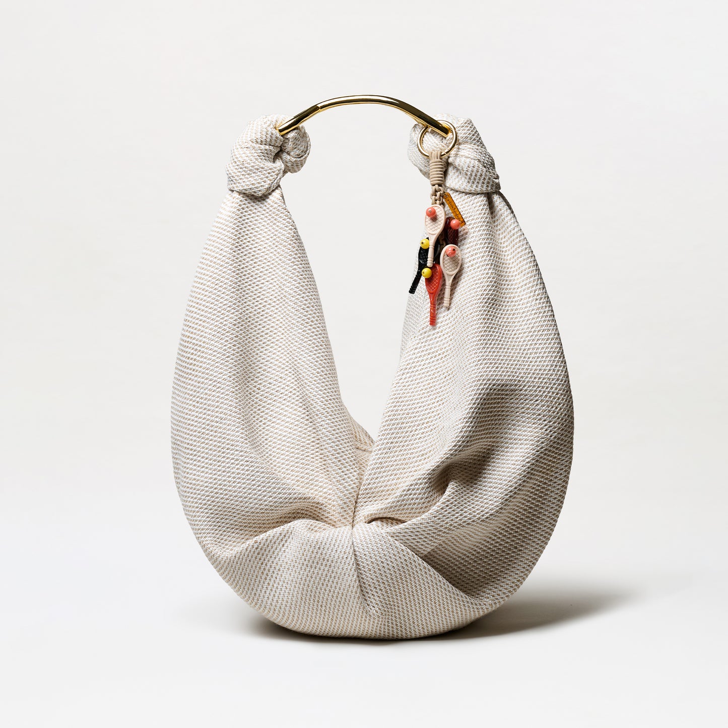 The Tournament Charm is a cream-colored, textured fabric hobo bag with a gold metal handle and several small, colorful keychain charms hanging from one side, displayed against a plain white background.