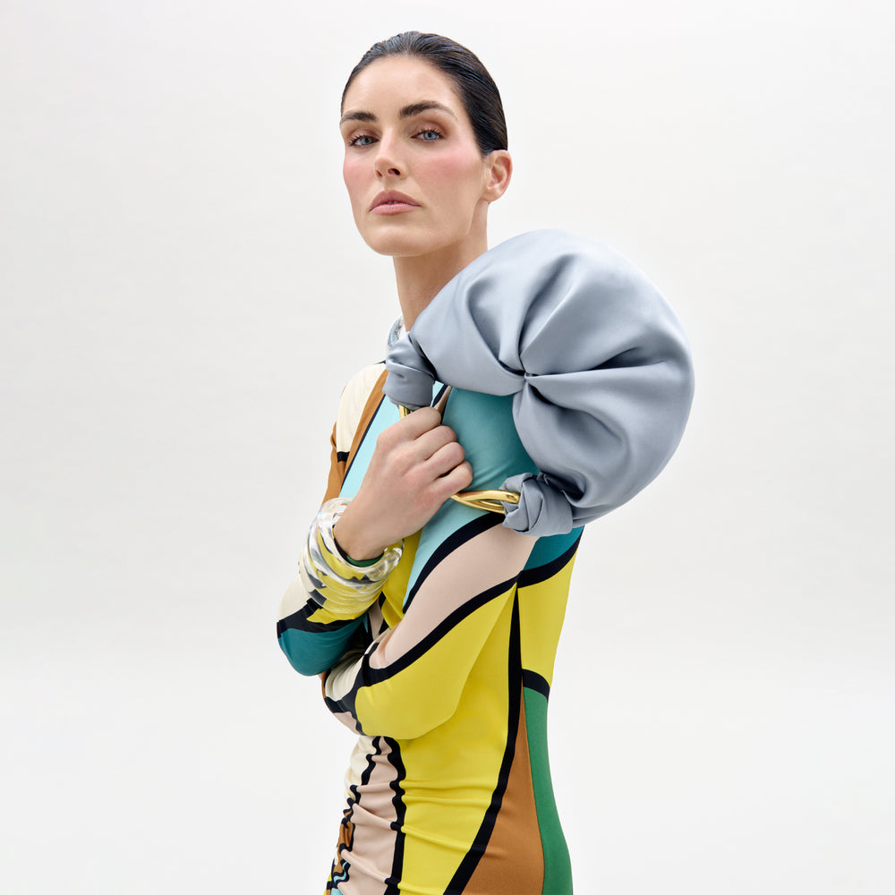 A woman in a colorful, geometric-patterned dress poses against a plain backdrop, holding the Sofia Bag (Small) in light blue on her shoulder. She wears bold makeup and chunky bracelets, gazing confidently at the camera.