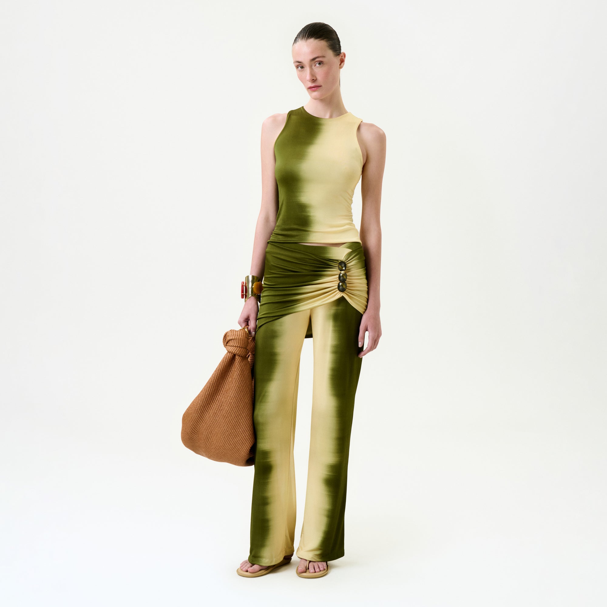 A person wearing the Sonu Pant, a slim fit green and yellow gradient pant with a side slit, holds a large brown textured bag while standing against a plain white background.