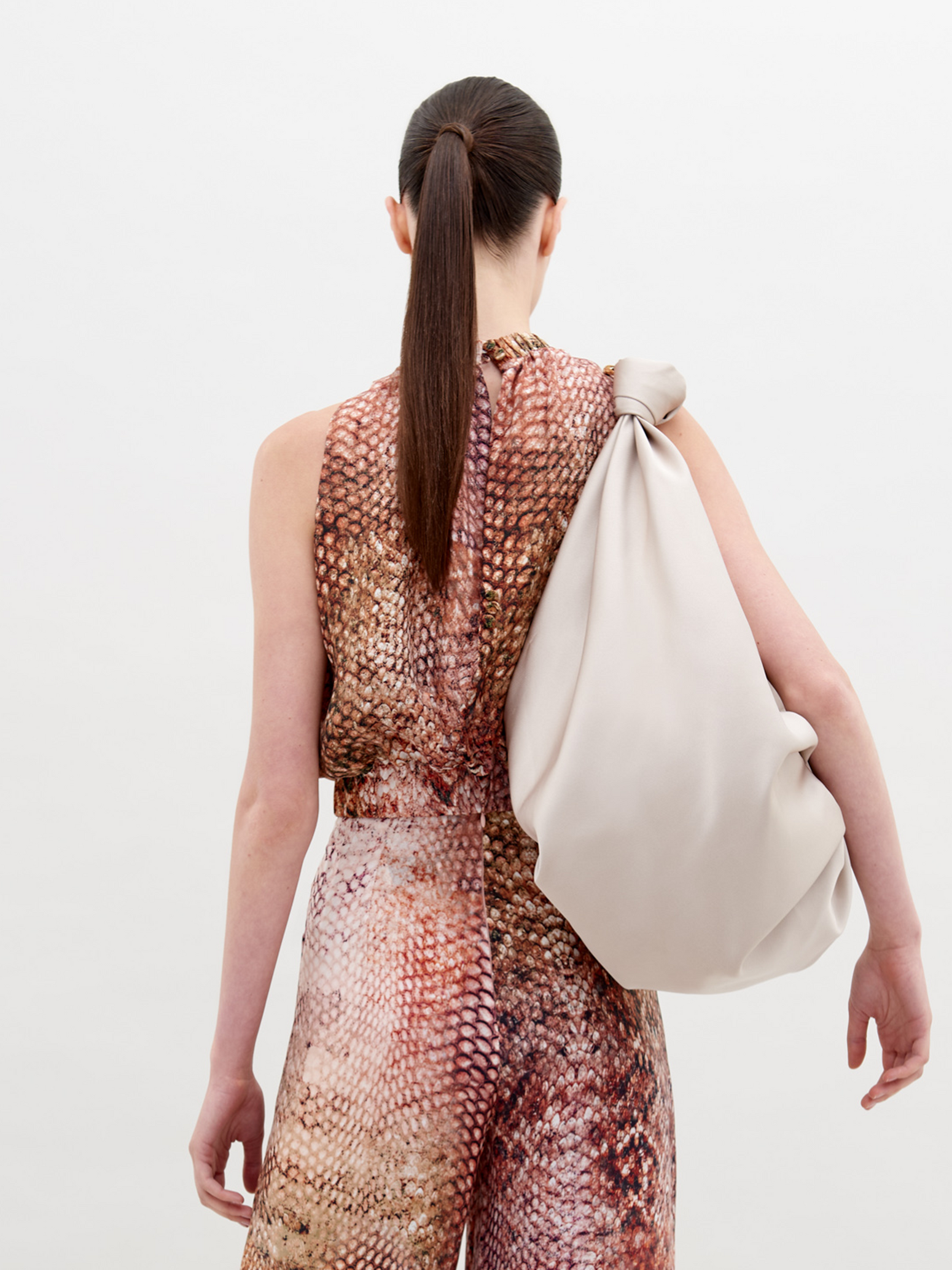 A person with tied-back hair wears a Stefano Blouse Peach Animal Print and matching pants, holding a soft, oversized beige bag on their shoulder against a plain white background. Available for pre-order now, shipping by November 15th, 2024.