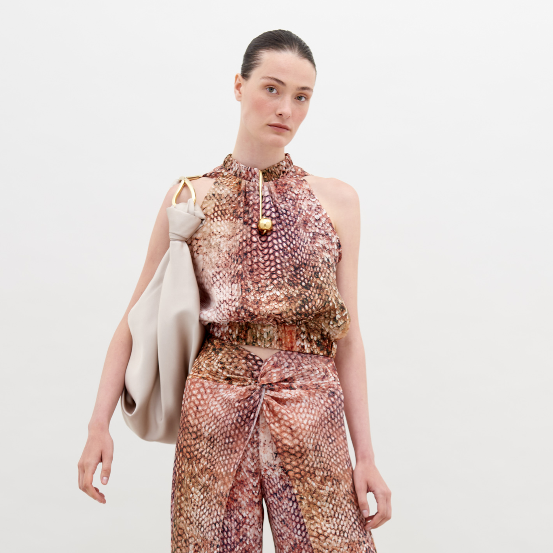 A person with tied-back hair wears a Stefano Blouse Peach Animal Print and matching pants, holding a soft, oversized beige bag on their shoulder against a plain white background. Available for pre-order now, shipping by November 15th, 2024.