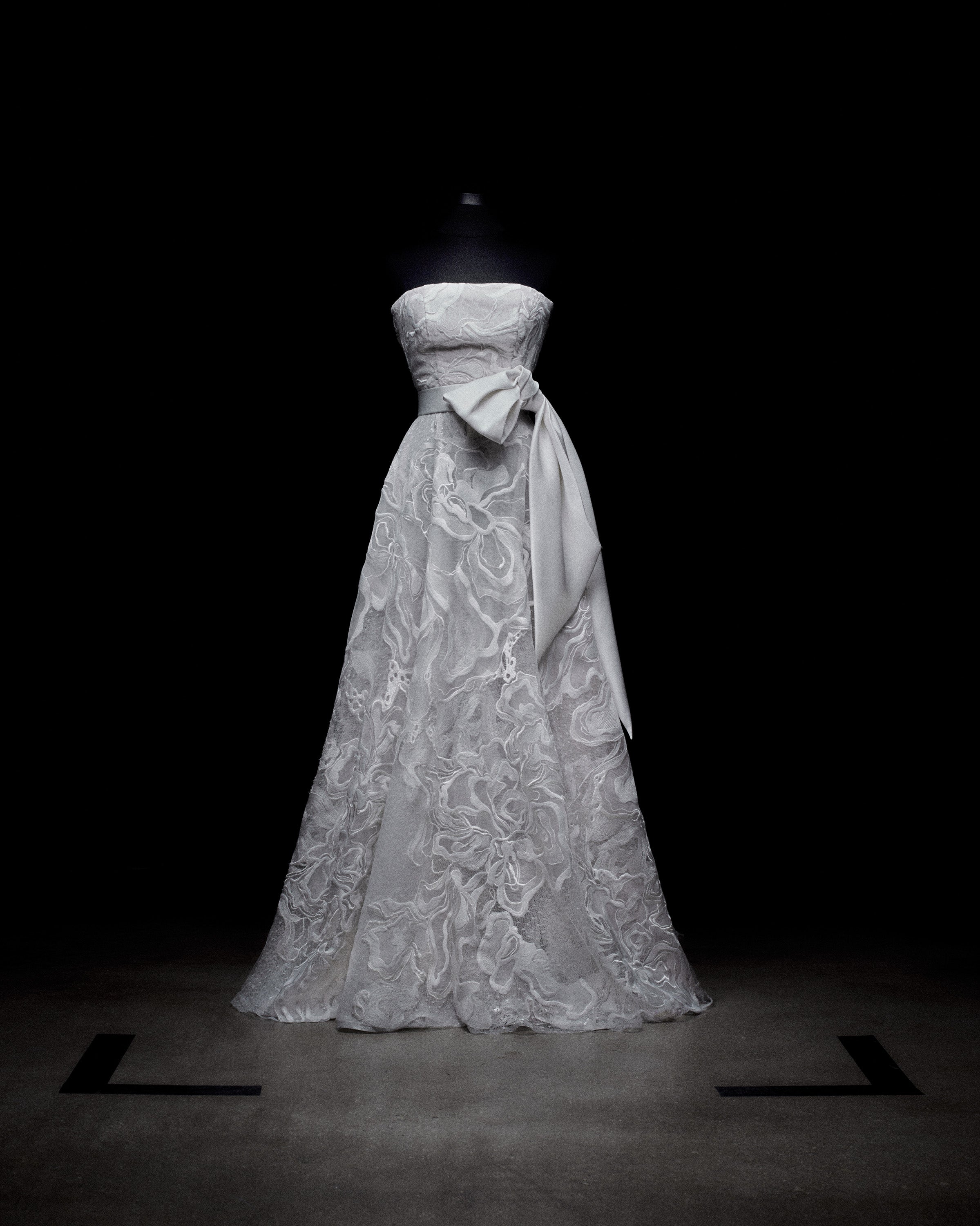 Strapless white gown on a mannequin, featuring intricate floral lace patterns and a large side bow, set against a dark background.