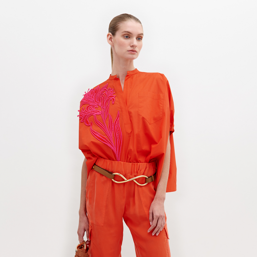A woman stands against a white background, wearing an orange outfit with a pink floral pattern on the upper left side and a gold belt. The Susanne Blouse Scarlet is available for pre-order until November 15th, 2024.