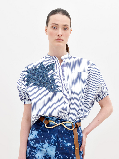 A person with straight, pulled-back hair wears a Susanne Blouse White Blue Stripes with a blue embroidered design on the front and blue patterned pants secured with a belt featuring a gold knot buckle. Pre-order now to ensure delivery by the ship date of November 15th.