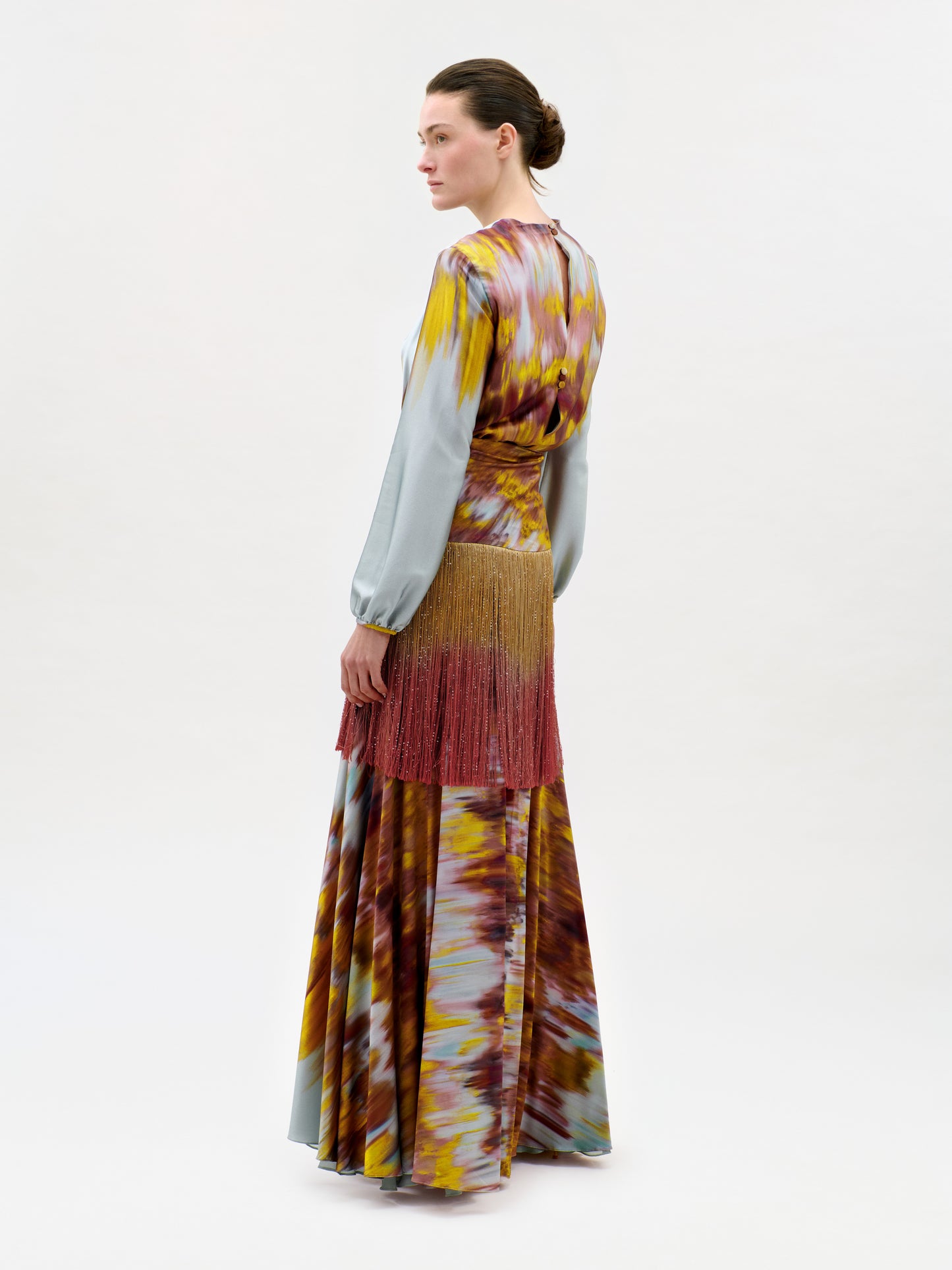 A woman stands sideways wearing the Sutton Dress, a long patterned maxi crafted in Colombia from luxurious Italian silk. The dress showcases shades of yellow, brown, red, and blue, with long sleeves and a distinctive fringed detail around the hips. Her hair is styled back in a neat bun.