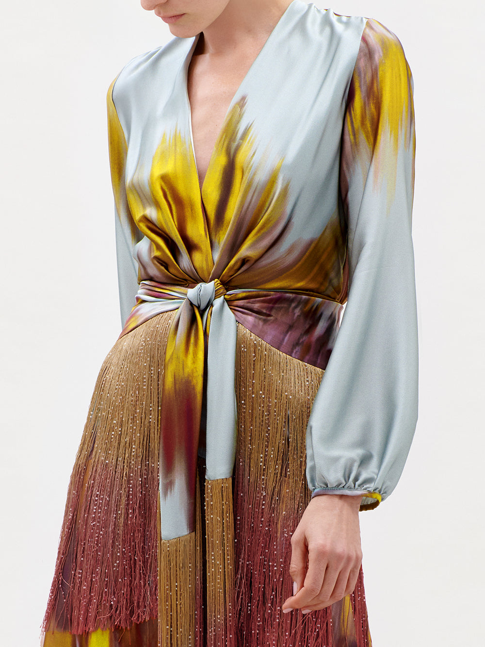 A woman wearing the Sutton Dress—a luxurious Italian silk maxi featuring long sleeves, a deep V-neckline, a knot at the waist, and a striking multicolored pattern in blue, yellow, and burgundy. This stunning garment is finished with fringe details along the skirt.