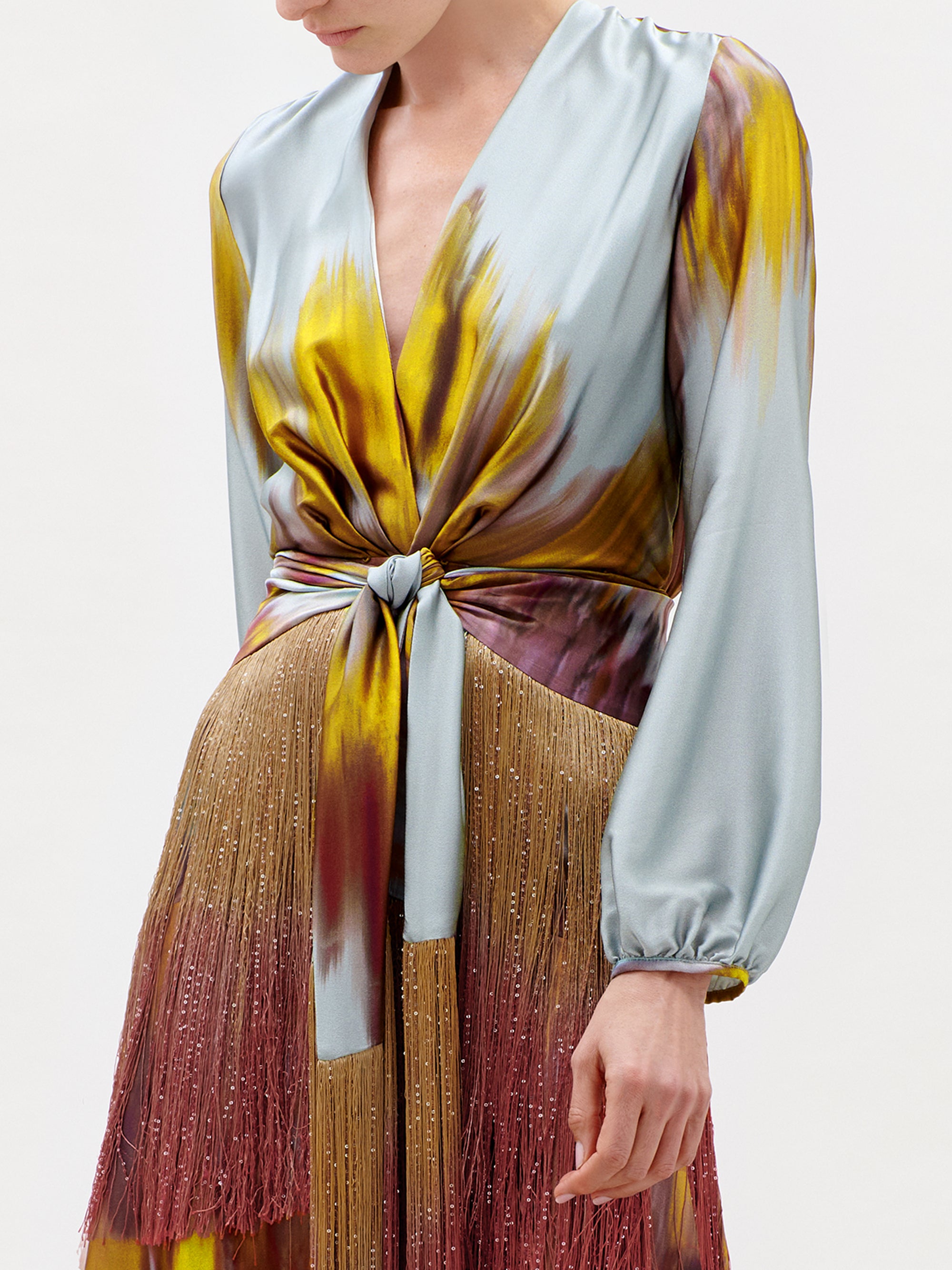 A woman wearing the Sutton Dress—a luxurious Italian silk maxi featuring long sleeves, a deep V-neckline, a knot at the waist, and a striking multicolored pattern in blue, yellow, and burgundy. This stunning garment is finished with fringe details along the skirt.