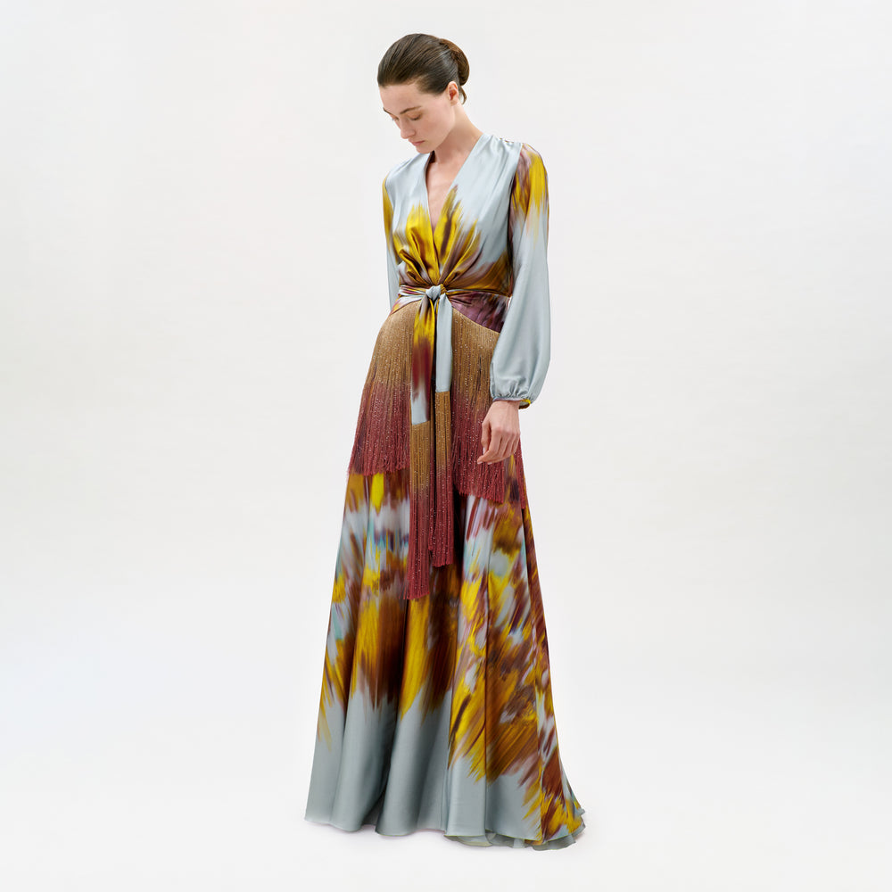 Wearing the Sutton Dress—a long, flowy silk maxi featuring a yellow, brown, and gray tie-dye pattern and crafted in Colombia—a person stands against a plain white background, looking down.