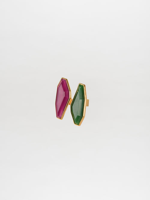 The Suzy Ring Magenta Emerald boasts a gold band adorned with two large, faceted gemstones in magenta and emerald hues, exuding a whimsical elegance.