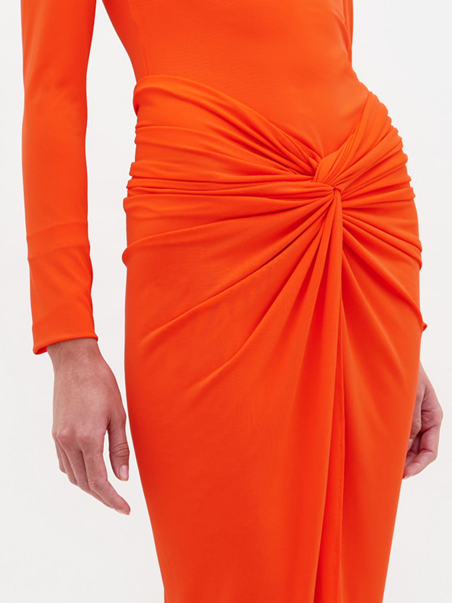 A person wearing the Sydelle Dress Tangerine, a long-sleeved orange dress featuring a knotted detail in the front, stands looking to the side. They are also donning sandals embellished with pearl-like beads. This elegant outfit will be available for pre-order and is anticipated to ship by 2025.