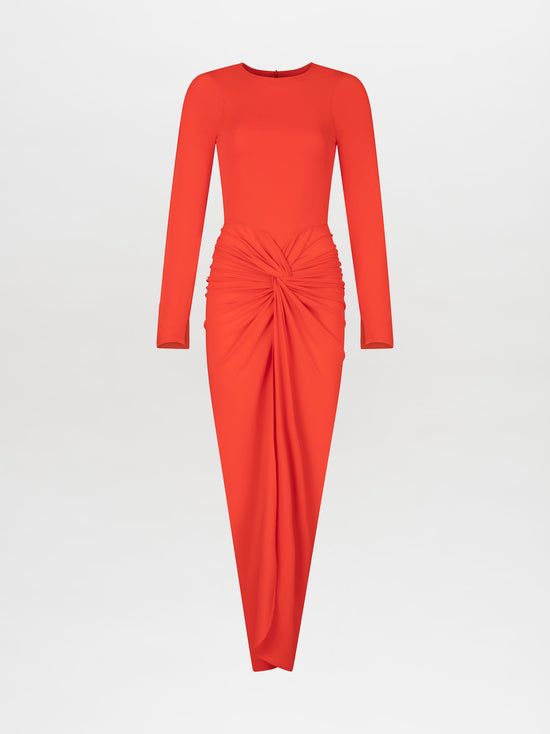 A person wearing the Sydelle Dress Tangerine, a long-sleeved orange dress featuring a knotted detail in the front, stands looking to the side. They are also donning sandals embellished with pearl-like beads. This elegant outfit will be available for pre-order and is anticipated to ship by 2025.