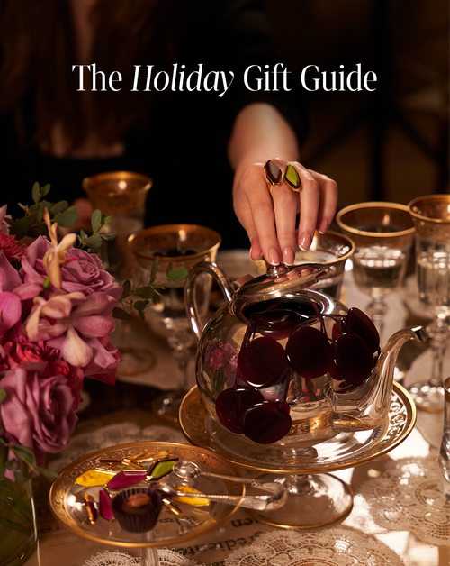 Chocolate pieces are carefully placed inside an ADA COMPLIANCE TEST PRODUCT teapot, creating an elegant table setting complete with flowers and gold-rimmed glassware. At the top, the words "The Holiday Gift Guide" are prominently displayed.