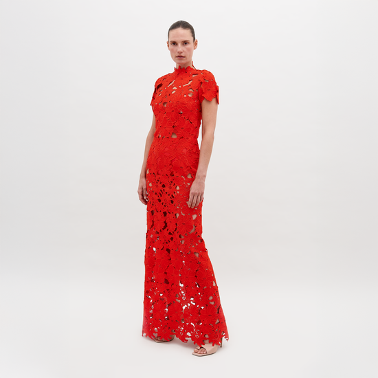 Person wearing a floor-length, red lace Taiana Dress Rouge with a high neckline and short sleeves, standing against a plain white background. Pre-order now to ensure delivery by the November 15th, 2024 ship-by date.