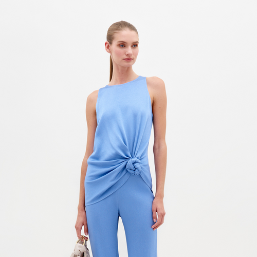 A person wearing the Tanima Blouse in periwinkle, a light blue sleeveless top with a knot at the waist, paired with matching pants and holding a small handbag, exudes effortless style. Perfect for pre-order, this chic ensemble ensures you'll be runway-ready by February 15th.