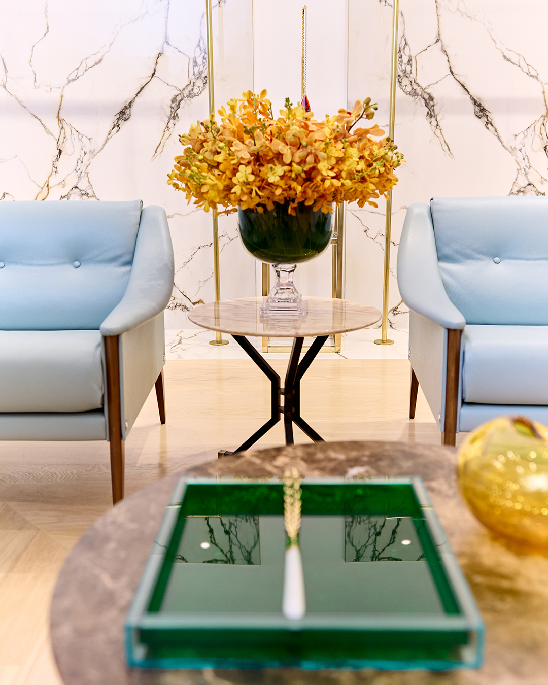 A modern living room with two blue armchairs, a round marble side table with a vase of orange flowers, and a large green tray on a central marble table. Marble wall in the background.