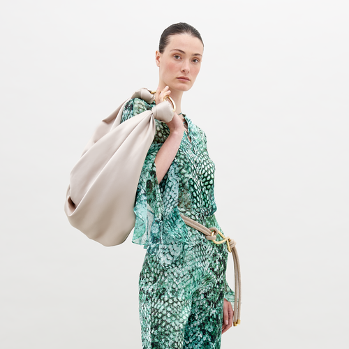 A person with slicked-back hair wears Thea Blouse Emerald Animal Print and holds a large beige bag over their shoulder, perfect for those looking to pre-order now. The background is plain white, highlighting the stylish ensemble expected to ship by November 15th, 2024.