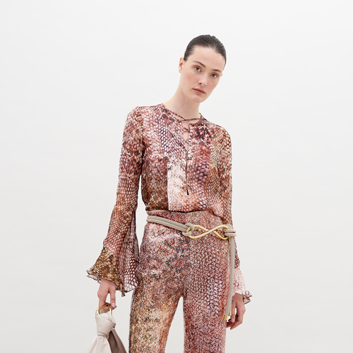 A person is wearing a Thea Blouse Peach Animal Print with flared cuffs and matching pants. They have a neutral expression and are holding a white accessory, available for pre-order now. Secure your piece before November 15th, 2024.