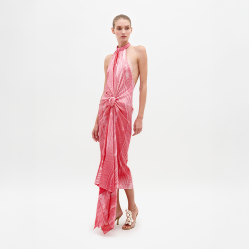 A person wearing Theodora Dress Peony Moire, a sleeveless, tie-dyed pink dress with a high neckline and front knot detail, paired with beige high-heeled sandals. Pre-order now to ensure you receive it by the ship-by date of February 15th, 2025.