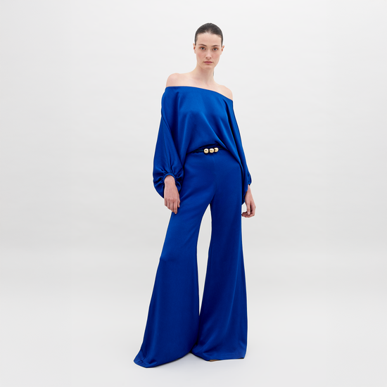 A person stands against a plain background wearing an off-the-shoulder blue top with wide sleeves and matching wide-leg pants, accessorized with a belt that has two large gold embellishments. Pre-order the Theresa Jumpsuit Royal Blue now to ensure you receive your outfit by the ship by date of November 15th, 2024.