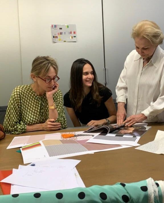 Three women are gathered around a table with design materials and papers. One woman stands, showing the others something in a magazine.