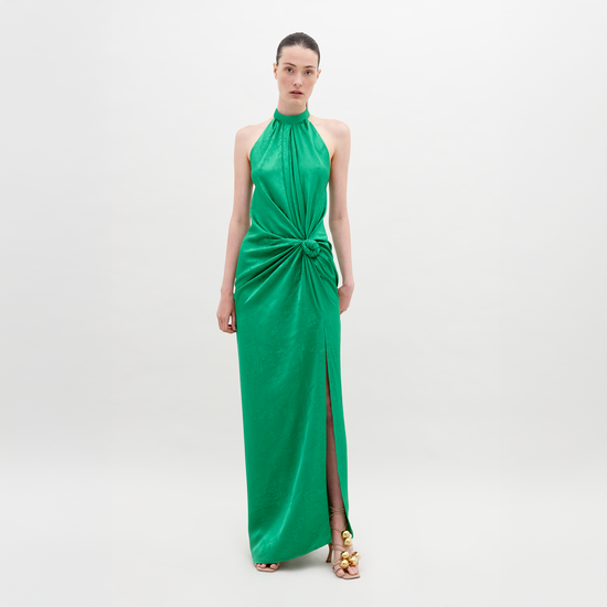 A woman stands against a plain background wearing a sleeveless, high-neck green dress with a front knot and a thigh-high slit. She has her hair pulled back and wears open-toed gold sandals. The Tizziana Dress Green is available for pre-order starting November 15th.