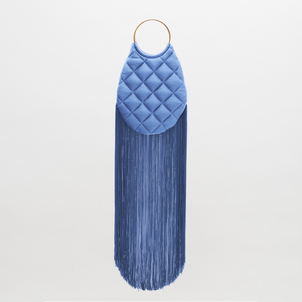 The Toblach Bag is a structured handbag in blue quilted Italian hammered satin, with a gold circular top handle and long, dense blue fringe at the bottom, shown against a plain white background.