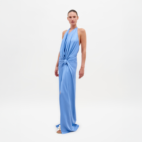 A person in the Torgiano Dress Periwinkle, a stunning blue halter-neck design with a central knot and an elongated hem, stands against a plain white background. This elegant piece is available for pre-order now and will ship by February 15th, 2025.