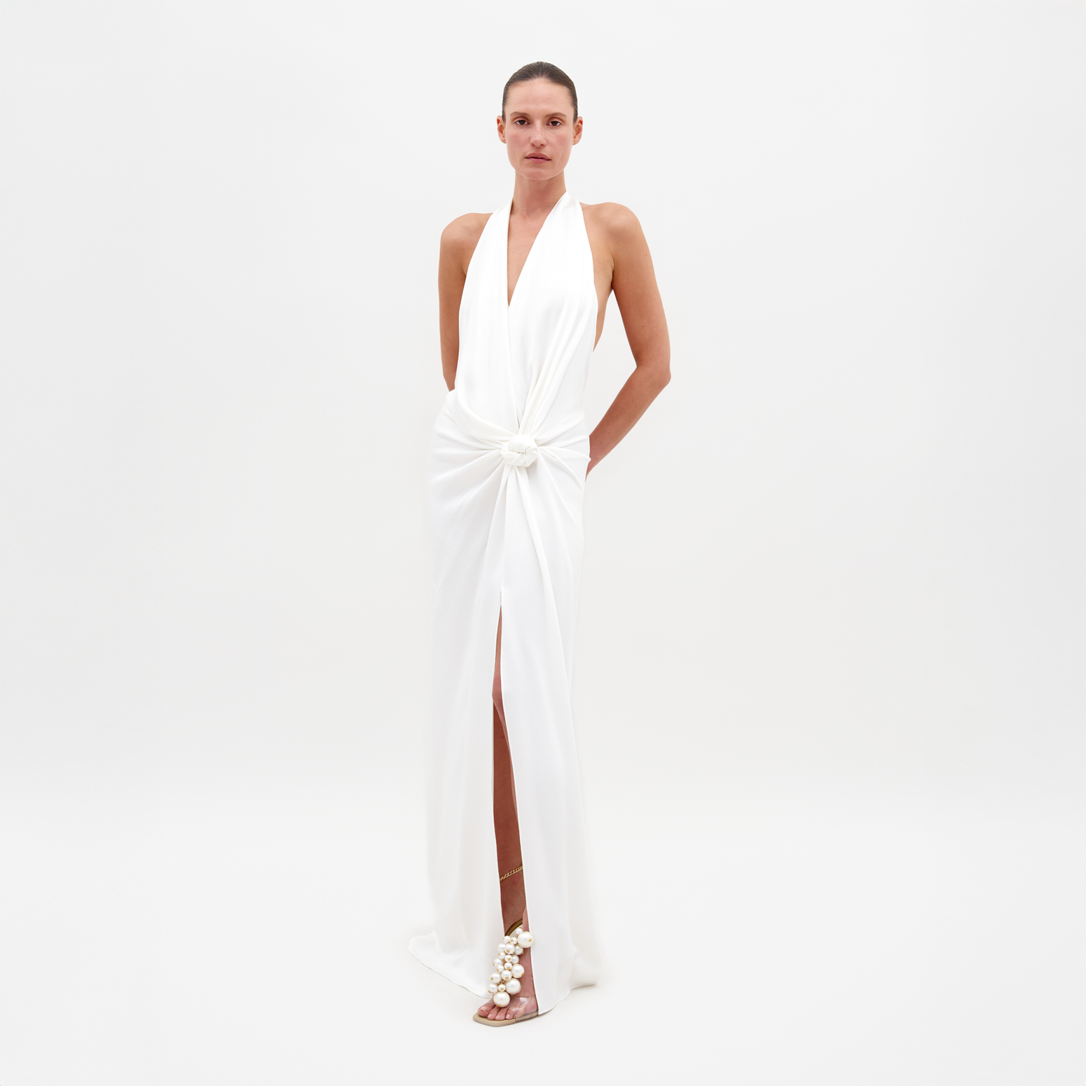 Person standing against a white background, wearing the Torgiano Dress White, which features a sleeveless halter design with a knot detail at the waist and a front slit. Pre-order now to ensure your dress is shipped by February 15th, 2025.